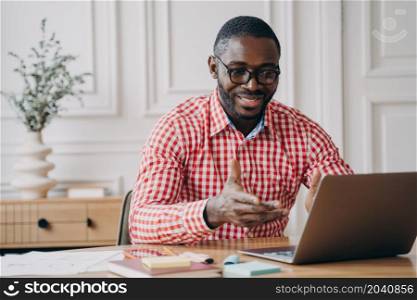 Positive african man online teacher tutor looking at laptop screen and smiling, talking with students while working remotely from home, dressed in casual clothes. E-learning and distant education. Positive african man online teacher tutor looking at laptop screen and talking with students