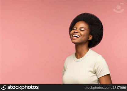Positive African woman in gray t-shirt happy laughing. Delightful black young adult girl cheerful smiling with closed eyes, isolated on pink studio background. Emotion of happiness and excitement.. Cheerful african woman happy laughing over pink wall. Delightful young girl smiling with closed eyes