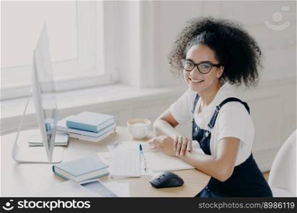 Positive Afro American female freelancer poses at workplace with papers and textbooks, works remotely on computer, has coffee break, works in her own cabinet, wears casual clothes. People, job