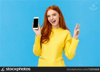 Positive and excited, charming redhead woman in yellow sweater, showing smartphone screen, advertise online store, mobile application, make peace sign and smiling joyfully, blue background.. Positive and excited, charming redhead woman in yellow sweater, showing smartphone screen, advertise online store, mobile application, make peace sign and smiling joyfully, blue background