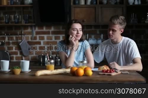 Positive attracitve couple spending leisure together and communicating while sitting at the kitchen table in the morning. Smiling affectionate man giving a kiss to his pretty girlfriend while having breakfast at home.