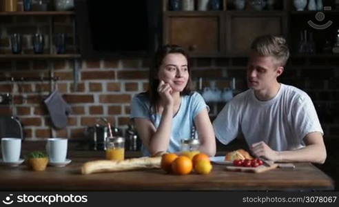 Positive attracitve couple spending leisure together and communicating while sitting at the kitchen table in the morning. Smiling affectionate man giving a kiss to his pretty girlfriend while having breakfast at home.