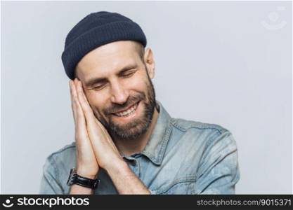 Positive bearded male pretends sleeping, has pleasant dreams, rests on hands, smiles joyfully, wears black stylish hat, isolated over white background. People, good rest and emotions concept.