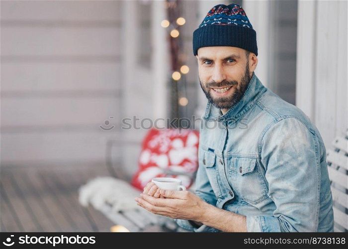 Positive bearded stylish male with pleasant smile, wears fashionable hat and jean jacket, holds cup of aromatic coffee, spends free time at balcony, breathes fresh air, thinks about something
