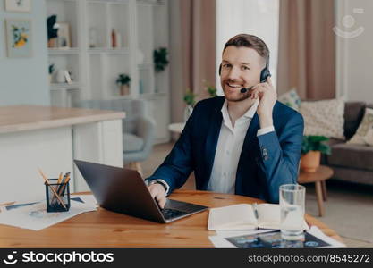 Positive bearded young man freelancer wearing suit in headphones with microphone smiling at camera while sitting at his workplace at home, using laptop for work and having online meeting. Smiling businessman in headphones sitting at desk at home office and working on laptop