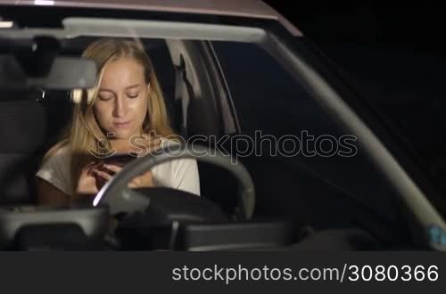 Positive beautiful long blonde hair female sitting in driver&acute;s seat and browsing the net with mobile phone on a summer night. Front view. Charming young woman in modern car using smart phone at night and smiling.