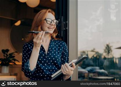 Positive beautiful woman with red hair holds phone talks on speakerphone with friend makes voice recognition or request uses internet services poses near cafeteria wears spectacles polka dot dress. Woman with red hair holds phone talks on speakerphone with friend makes voice recognition