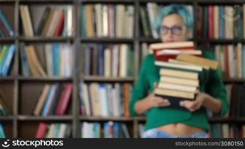 Positive beautiful young woman with blue hair and trendy spectacles carrying a stack of books in university library. Serious college female student preparing for exams, doing research in library with pile of books. Midsection.