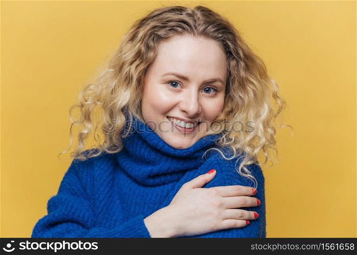 Positive blonde curly young woman with broad smile, dressed in casual blue sweater, smiles gently, isolated over yellow studio background. Delighted female student passed final exam successfully