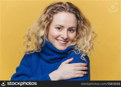 Positive blonde curly young woman with broad smile, dressed in casual blue sweater, smiles gently, isolated over yellow studio background. Delighted female student passed final exam successfully