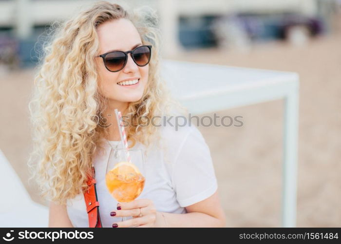 Positive blonde female with curly hair, wears sunglasses, looks with happy expression aside, has gentle smile, holds fresh cocktail, waits for friend on beach. People, resort, recreation concept