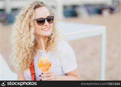 Positive blonde female with curly hair, wears sunglasses, looks with happy expression aside, has gentle smile, holds fresh cocktail, waits for friend on beach. People, resort, recreation concept
