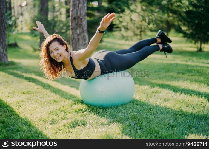 Positive brunette leads active life with fitness ball, posing in black clothes against forest backdrop. Happy smile, preps for Pilates workout.