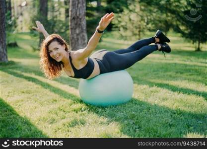 Positive brunette leads active life with fitness ball, posing in black clothes against forest backdrop. Happy smile, preps for Pilates workout.