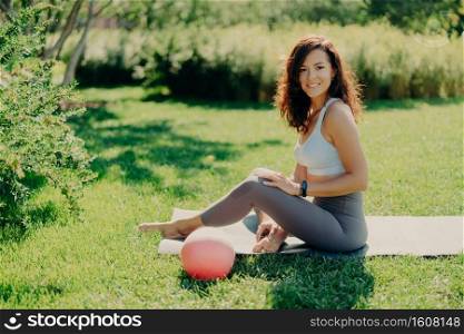 Positive brunette woman in cropped top and leggings poses bare feet on karemat uses ball for gymnastic exercises smiles pleasantly enjoys beautiful nature view. Sport outdoor. Yoga practice.