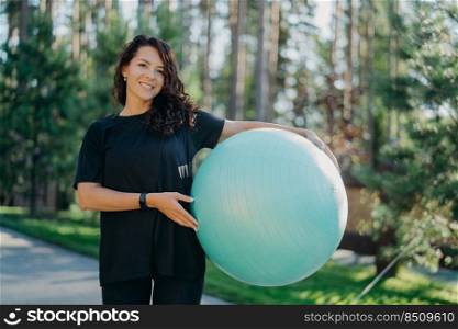Positive brunette woman leads active lifestyle holds fitness ball poses against forest background dressed in black clothes has happy smile. Pretty female prepares for workout pilates does exercises