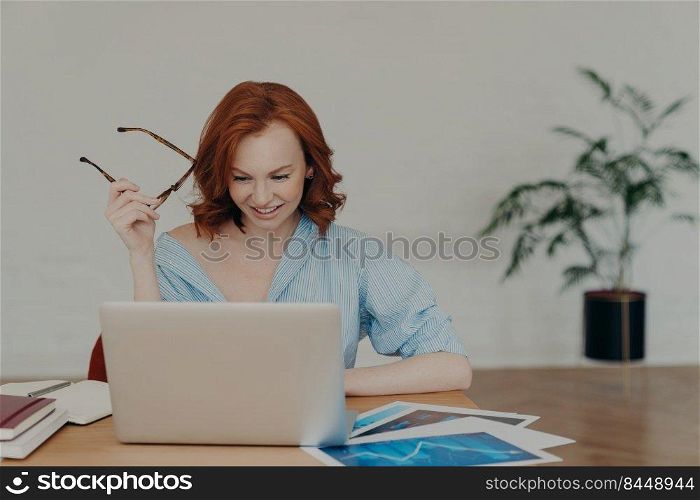 Positive busy woman with European appearance works on freelance digital project, earns money online on distance job, sits at workplace with papers and notepad, wears casual shirt, holds glasses
