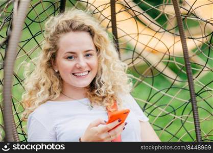 Positive Caucasian female with curly hair, sits in hanging chair, has friendly smile, types text messages on modern cell phone, makes shopping online, enjoys summer time and unforgettable rest