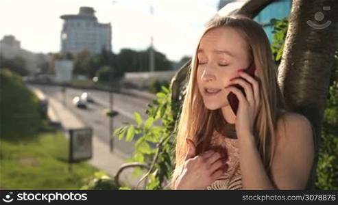 Positive charming blond woman talking on mobile phone over cityscape background. Smiling blond girl comminicating on smartphone while leaning on the tree in the park at sunset. Slow motion. Stedicam stabilized shot.