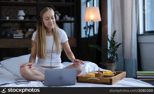 Positive charming long blonde hair female eating healthy breakfast while sitting crossed legs on white bed sheet early morning over modern domestic background. Beautiful young woman in sexy white underwear relaxing at home during morning breakfast.