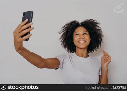 Positive cheerful dark skinned female making photo on modern smartphone camera, cheerful african woman dressed in casual white tshirt taking selfie on her mobile phone, smiling happily. Positive cheerful dark skinned female making photo on modern smartphone camera