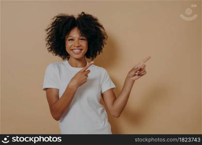 Positive cheerful dark skinned woman dressed in white basic tshirt pointing at copy space isolated on beige background, advertising displaying product, showing place for your advertisement promotion. Positive cheerful dark skinned woman pointing at copy space isolated on beige background