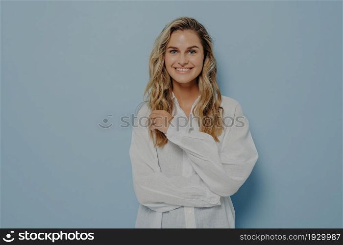 Positive cheerful young beautiful woman with fair wavy hair in casual clothes smiling sincerely at camera, posing with hands folded, one hand touching hair, isolated on blue background with copy space. Cheerful young beautiful woman smiling sincerely at camera