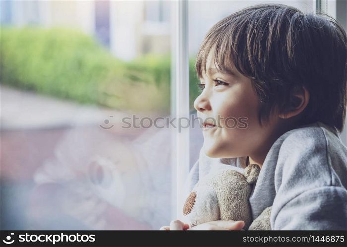 Positive child boy sitting at the window writing on glass and looking out with smiling face, School kid self isolation relaxing at home during qualrantine covid 19, Home schooling , Distance education