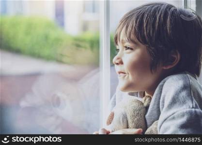 Positive child boy sitting at the window writing on glass and looking out with smiling face, School kid self isolation relaxing at home during qualrantine covid 19, Home schooling , Distance education