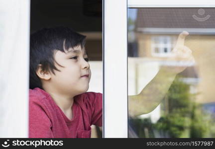 Positive child boy sitting at the window writing on glass with smiling face, School kid self isolation relaxing at home during covid 19, Home schooling , Distance education