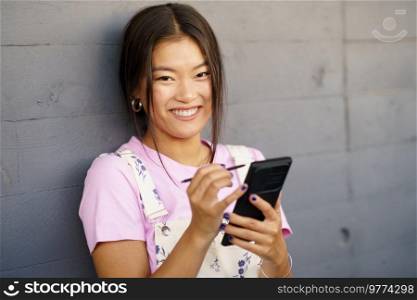 Positive Chinese woman using her smartphone with a pen or stylus, looking at camera outdoors. Asian female wearing casual clothes.. Positive Chinese woman using her smartphone with a pen or stylus, looking at camera outdoors