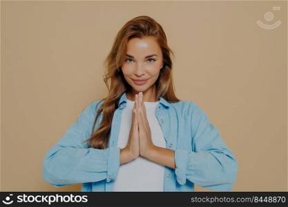 Positive concentrated young woman holding palms together in prayer gesture, being calm and peaceful, making namaste yoga sign on beige background. Body language concept. Young woman making namaste gesture isolated on beige background