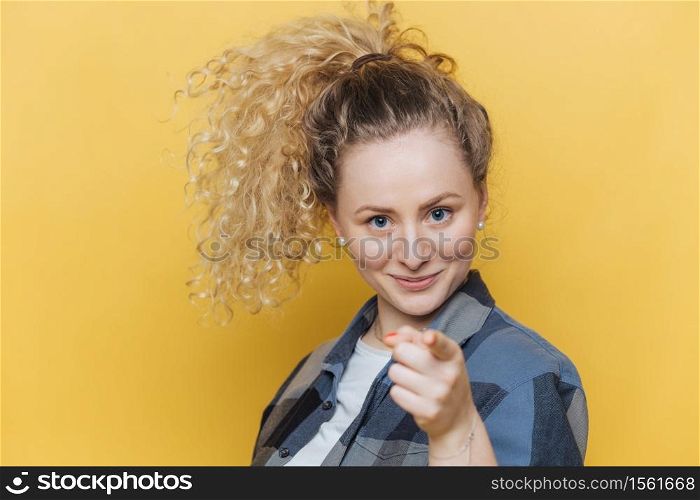 Positive confident beautiful female with curly pony tail, indicates directly at camera, chooses you, dressed in casual clothing, isolated over yellow background. Shot of pretty woman gestures indoor