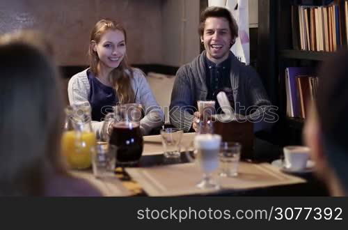 Positive couple of college students drinking coffee together in cafe while communicating with their friends. Group of hipster teenagers relaxing, laughing and having fun at the restaurant after lessons.