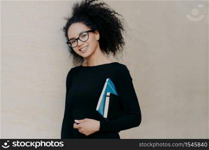 Positive curly African American student carries notepad and diary, wears black clothes, transparent glasses, enjoys studying in university, isolated on grey background. Cute woman with notebook