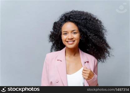 Positive curly dark skinned woman with luminous hair, wears formal purple jacket, poses against grey background with blank space, happy to meet with business partners. Emotions. Happy director