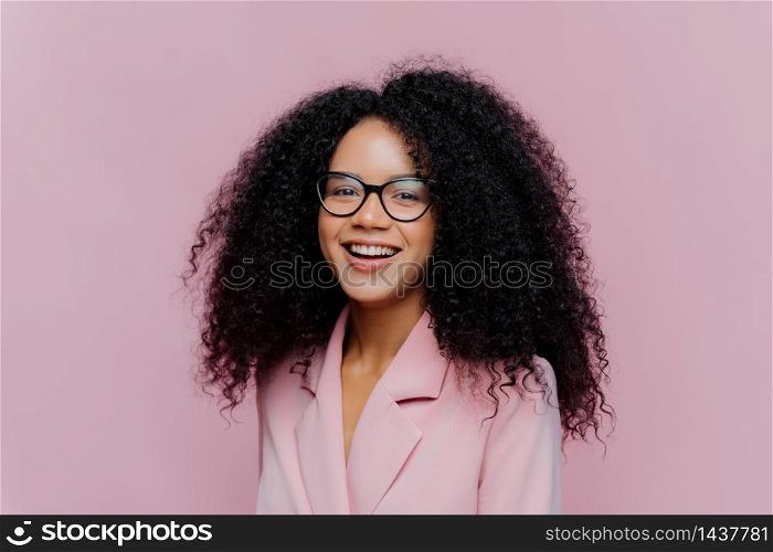 Positive curly haired businesswoman wears spectacles, formal wear, has bushy hairstyle, being in good mood after successful day at work, poses against violet background. Monochrome. Happy director