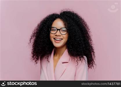 Positive curly haired businesswoman wears spectacles, formal wear, has bushy hairstyle, being in good mood after successful day at work, poses against violet background. Monochrome. Happy director