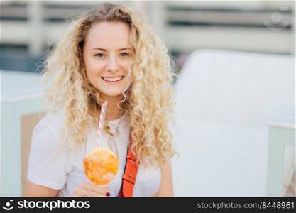 Positive curly young female with healthy clean skin, dressed in casual white t shirt, holds cold beverage, looks happily at camera, enjoys summer rest, breathes fresh marine air outdoor at beach