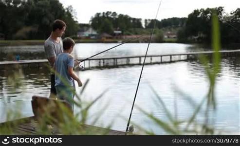 Positive dad and son with fishing rod angling at freshwater pond while standing on wooden pontoon and talking. Happy father and teenage boy spending great time together fishing on the lake in summer against amazing rural landscape background.