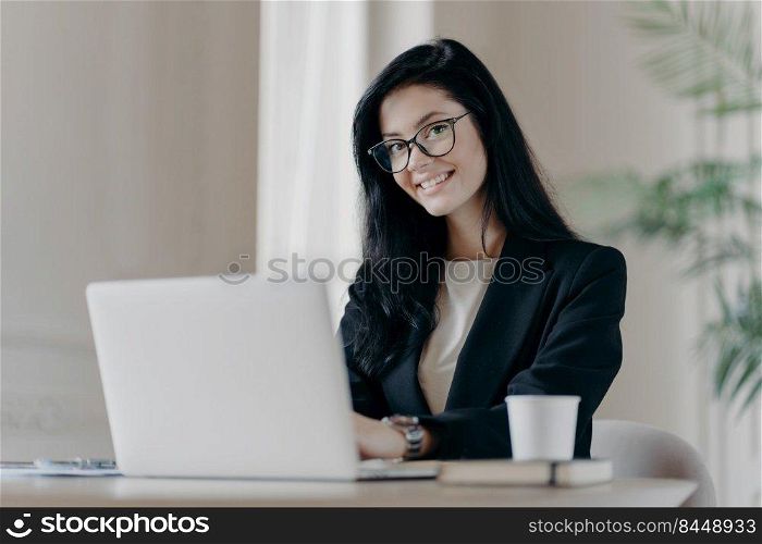 Positive dark haired woman makes research, browses information on laptop computer, connected to wireless internet, being happy wears formal clothes and spectacles drinks coffee. Economist works online