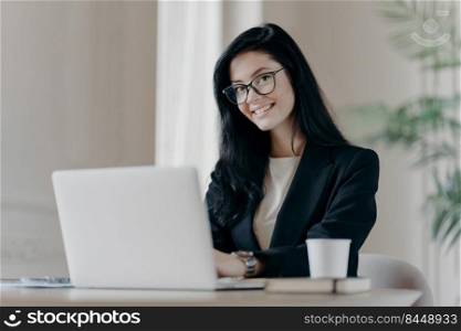 Positive dark haired woman makes research, browses information on laptop computer, connected to wireless internet, being happy wears formal clothes and spectacles drinks coffee. Economist works online
