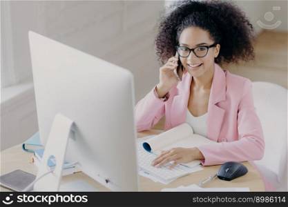 Positive dark skinned woman with curly combed hair, keybaords on computer, involved in working process, wears spectacles and elegant clothing, has telephone conversation, poses at workplace.