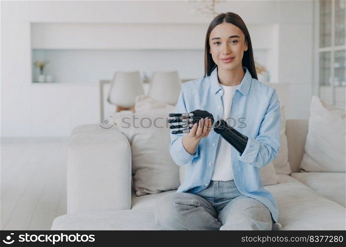 Positive disabled young woman sitting and disassembling bionic arm prosthesis. Cyber sensor artificial arm has processor chip and buttons. European girl has high tech carbon robotic hand and smiling.. Positive disabled young woman is disassembling bionic arm prosthesis and smiling.
