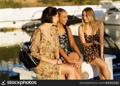 Positive diverse female friends in dresses chatting while sitting on wharf near river with moored boats on sunny summer day. Cheerful multiethnic women sitting on pier