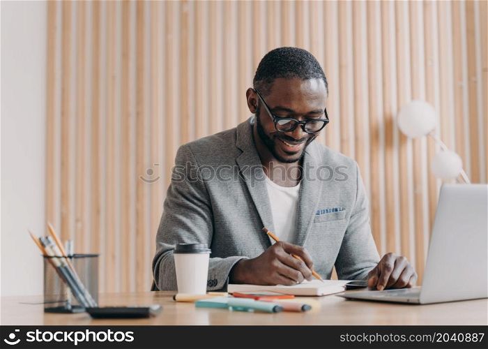 Positive elegant african male employee having video conference online with colleagues, smiling american businessman makes notes in agenda while sits at desk at home office and using laptop. Happy afro american smiling businessman makes notes in agenda while having video call online