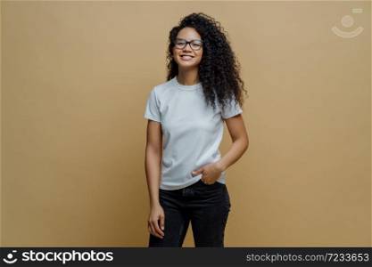 Positive ethnic female student feels free after passing exam, smiles broadly, keeps hand in pocket of jeans, wears transparent glasses, poses against brown background. People, lifestyle concept