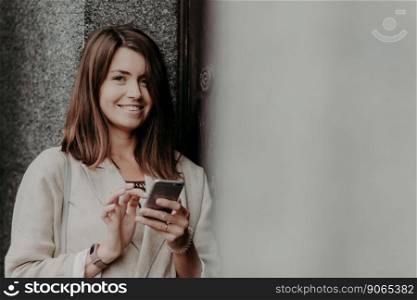 Positive European female with pleasant appearance, reads income message, watches video online, chatts in social networks, uses mobile phone, has happy expression. People, emotions, lifestyle concept