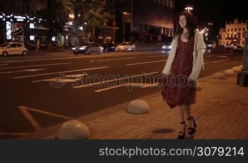 Positive fashionable woman in stylish clothes and high heels walking along city street alone at night over traffic and urbanscape background. Cheerful brunette girl strolling cobblestone street in the evening and smiling. Steadicam stabilized shot.