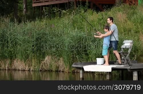 Positive father helping his teenage boy to fight a fish they caught. They are spinning fishing reel while fishing together from wooden pier on the pond. Cute son and dad catching fish with fishing rod and reel while spending leisure on the lake.